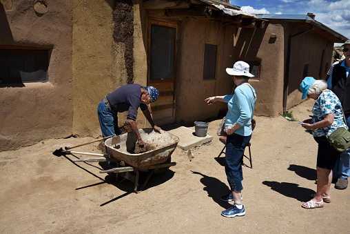 TAOS, NEW MEXICO - MAY 15, 2019: Tourists watch as a Native-American man repairs his adobe home at Taos Pueblo, an ancient Native-American pueblo near Taos, New Mexico USA.