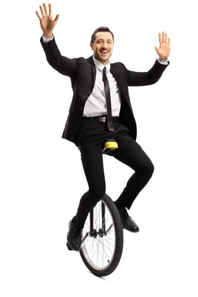 istock Young businessman in a suit riding a unicycle and waving with both hands 1158507674