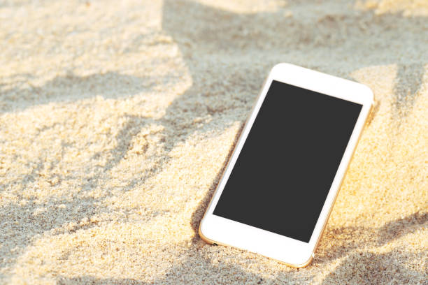 istock mobile smart phone put drop on tropical sand beach with sunlight texture background. display black copy space of technology. Forgot to fall lost on the way 1158518154