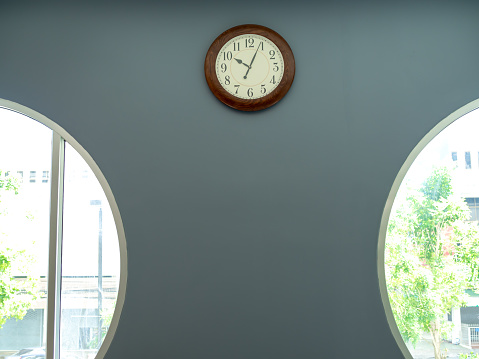 Round vintage wooden wall clock hanging on the grey wall near round window with copy space.