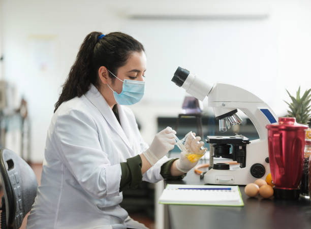 Female lab student wearing protection for experiment A latin female lab student wearing gloves and mask, sitting and holding a flask with an egg inside. egg food photos stock pictures, royalty-free photos & images