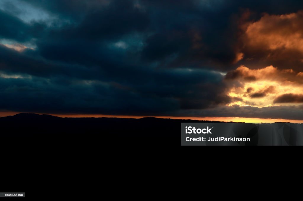 Cloudy Rural Sunset with Hills and Trees Silhouetted A country sunset with sunlight glowing through the clouds at right and highlighting the turquoise colors of the storm cloud. Hill and trees are silhouetted. Photographed at Maleny, Queensland, Australia. Australia Stock Photo