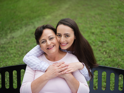 A portrait of a beautiful young woman standing behind her mother embracing her as they both look at the camera and smile.