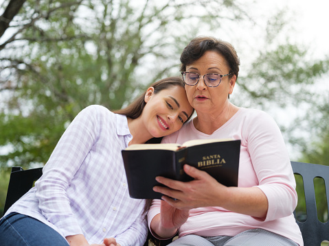 A young woman resting her head on her mothers shoulder as she reads to her from the Bible while sitting outdoors.