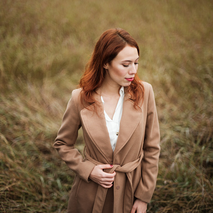 A portrait of a beautiful young woman standing in a meadow of brown grass with a brown trench coat while looking away from the camera.
