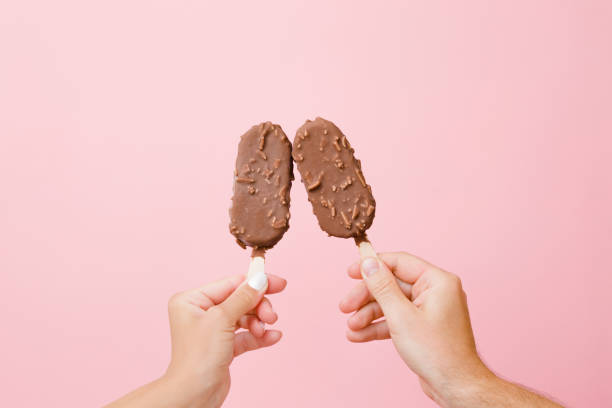 Young couple hands holding together ice creams with nuts and chocolate glaze on pink background. Pastel color. Close up. Point of view shot. Young couple hands holding together ice creams with nuts and chocolate glaze on pink background. Pastel color. Close up. Point of view shot. flavored ice photos stock pictures, royalty-free photos & images