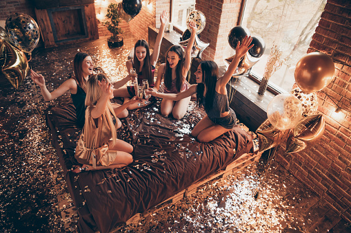 Photo of five diverse attractive pretty excited raising gesturing hands up ladies saying dreams, wishes to birthday girl in golden apron sitting on sheet linen decorated indoors loft interior