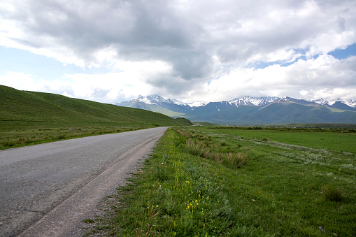 Country road among green fields and hills against the backdrop of mountains with snow-capped peaks and a cloudy sky. Traveling in Kyrgyzstan