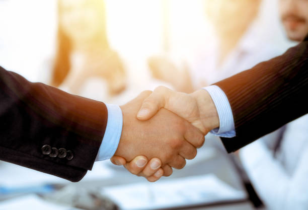 istock Business people shaking hands at meeting while theirs colleagues clapping and applausing. Group of unknown businessmen and women in modern white office. Success teamwork, partnership and handshake concept. 1158602783
