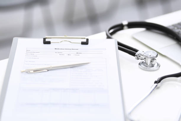 istock Stethoscope, clipboard with medical form lying on hospital reception desk with laptop computer and busy doctor and patient communicating at the background. Medical tools at doctor working table.Medicine concept. 1158602870
