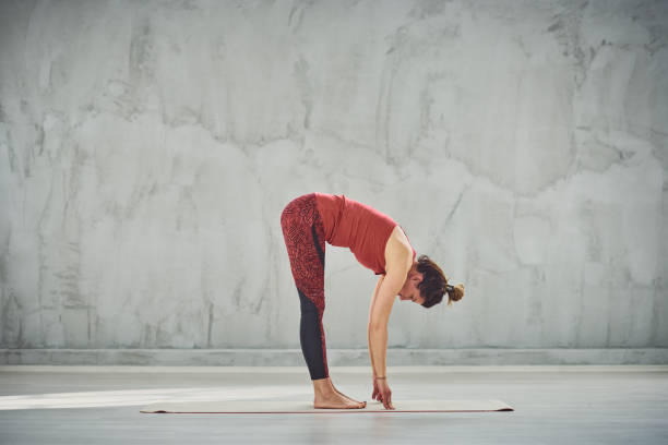 Woman practicing yoga. Side view of beautiful Caucasian brunette in red sports wear standing barefoot on the mat in Standing Forward Bend yoga posture. bending stock pictures, royalty-free photos & images