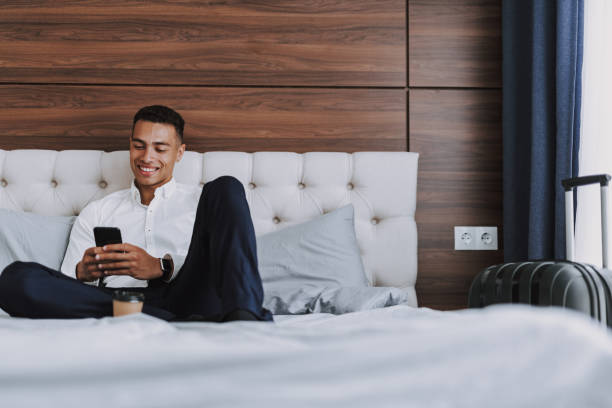 l'uomo allegro con lo smartphone sta bevendo caffè in camera da letto - hotel wireless technology bedroom hotel room foto e immagini stock