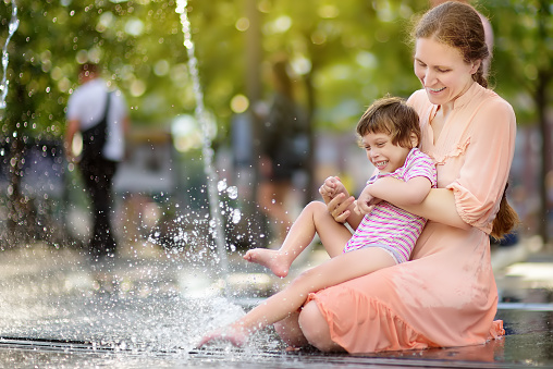 Portrait of beautiful disabled girl in the arms of his mother having fun in fountain of public park at sunny summer day. Child cerebral palsy. Inclusion.