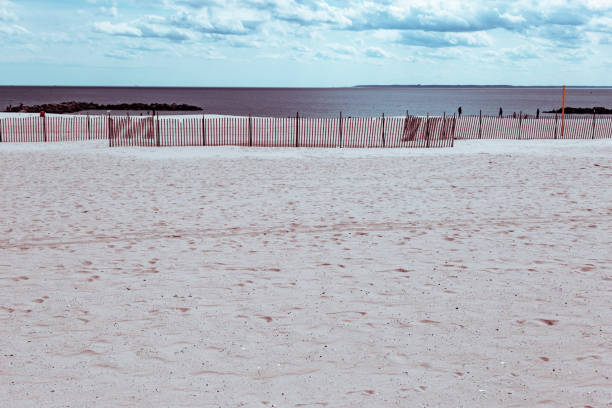 A black and white photograph of a beach with a white fence in the foreground and a dramatic sky filled with clouds in the background