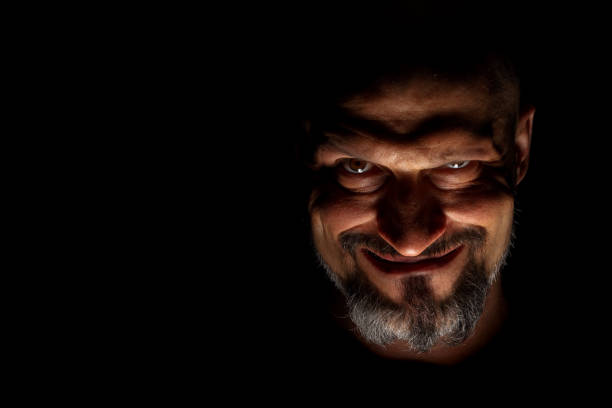 istock Face with a bearded man grimace against a dark background with sharp shadows. Comedic, fabulous villain or negative character conception with copy space. 1158613821