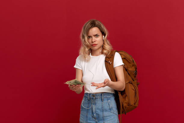 retrato de una chica con el pelo rubio rizado, mochila naranja y auriculares blancos vestidos con una camiseta blanca de pie sobre un fondo rojo que expresa emoción desconcertada porque no tiene suficiente dinero. - blowing hair audio fotografías e imágenes de stock