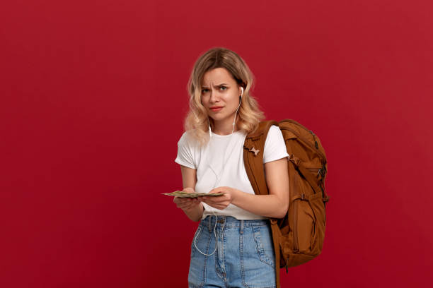 retrato de una chica con el pelo rubio rizado, mochila naranja y auriculares blancos vestidos con una camiseta blanca de pie sobre un fondo rojo que expresa emoción desconcertada porque no tiene suficiente dinero. - blowing hair audio fotografías e imágenes de stock