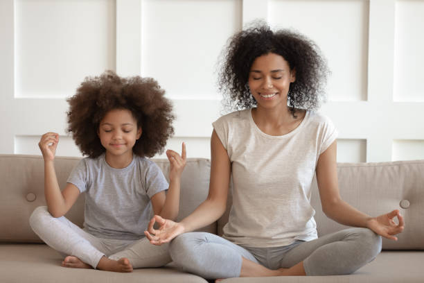 Healthy african mother teaching meditation sit with daughter on sofa Healthy calm african american mother teaching meditation sit with cute small child daughter on sofa, mindful happy black mom and kid girl doing yoga exercises relaxing together in lotus pose at home mindfulness kids stock pictures, royalty-free photos & images