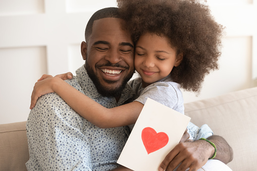 Happy affectionate african american dad embracing little child daughter holding greeting card with red heart bonding on fathers day concept, smiling cute kid girl hug daddy congratulate make surprise