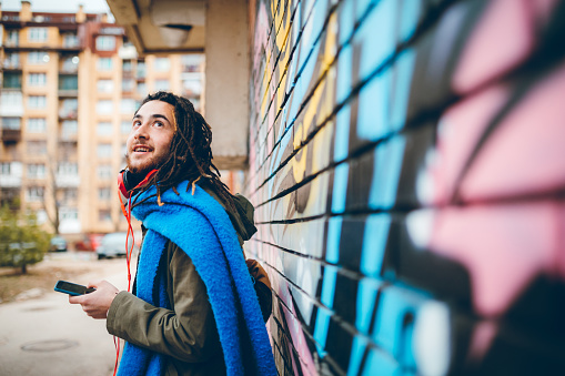 Young caucasian man with dreadlocks enjoys music leaning on a graffiti.