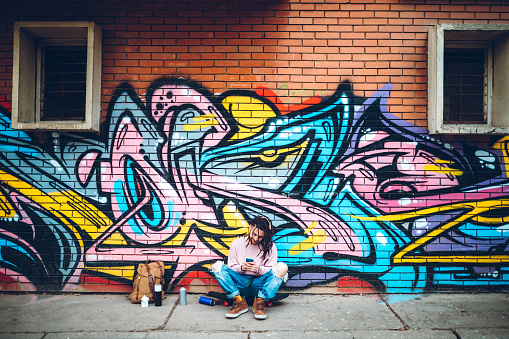 Young caucasian graffiti artist using phone and sitting on a skateboard.