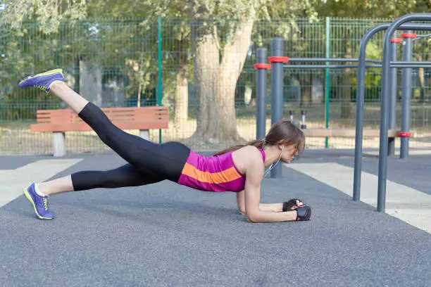 Young caucasian woman workouts on the park sportsground. Slim girl in plank position, one leg up, bright sportswear. Earphones, protective gloves. Woman morning training. Outdoor, copy space.