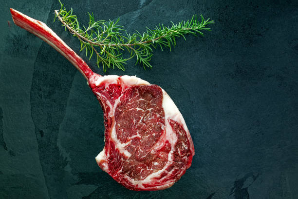 Raw, dry-aged wagyu tomahawk steak, salt,pepper, bbq rub and rosemary on a  black stone table. Top view stock photo