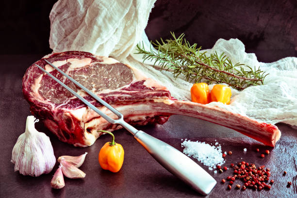 Raw ,dry-aged wagyu tomahawk steak with rosemary, salt, pepper and a meat fork on a black stone table. Front view