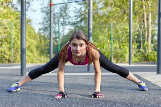 Young caucasian woman workouts on the park sportsground. Active strenght and stretching, bright sportswear. White earphones, protective gloves. Woman morning training, ironic look. Outdoors.
