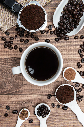 Direct above view of roasted coffee beans, coffee types in spoons,  coffee cup with coffee and coffee maker on wooden background.