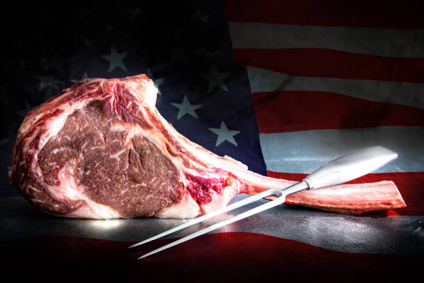 4th of July. Raw, dry aged Wagyu Tomahawk steak and meat fork with american flag in the background