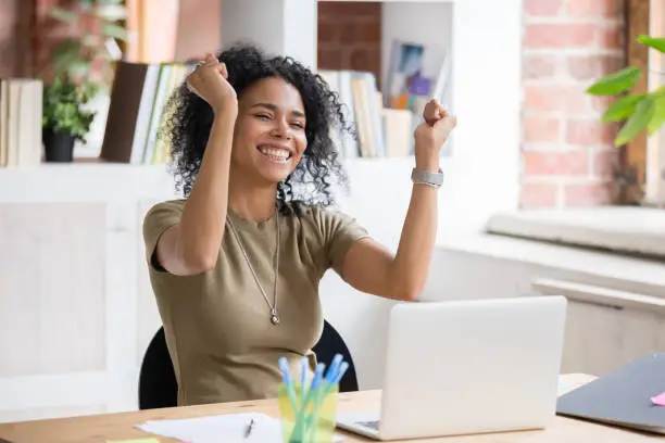 Excited african American woman sit at desk feel euphoric win online lottery, happy black woman overjoyed get mail at laptop being promoted at work, biracial girl amazed read good news at computer