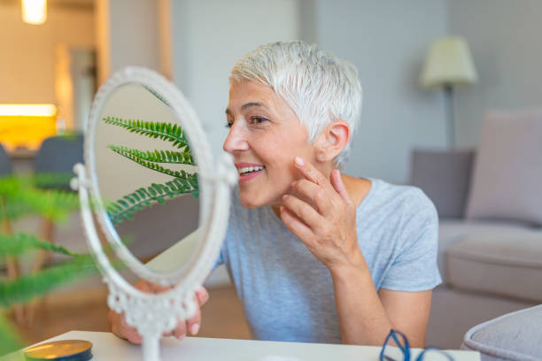 Beautiful elderly woman holding mirror and applying face cream at home Woman Looking At Herself In The Mirror. Beautiful elderly woman holding mirror and applying face cream at home. Senior woman applying anti-wrinkles cream. vanity mirror photos stock pictures, royalty-free photos & images
