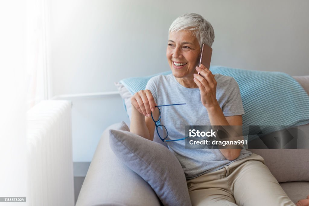 Senior woman talking on her mobile phone. Senior woman talking on her mobile phone. Senior woman has a happy conversation at cellphone. Smiling senior woman using phone sitting on couch at home. Using Phone Stock Photo