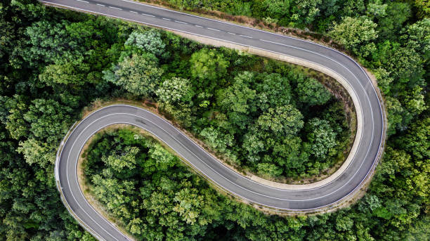 Winding road seen from above, with two hairpin bends, surrounded by a green forest with lush foliage. Aerial view, no people.