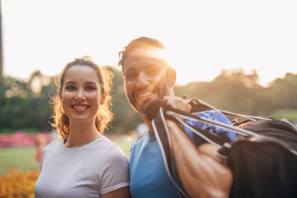 One young couple in sport clothing standing at park holding gym bag, looking at camera and smiling.