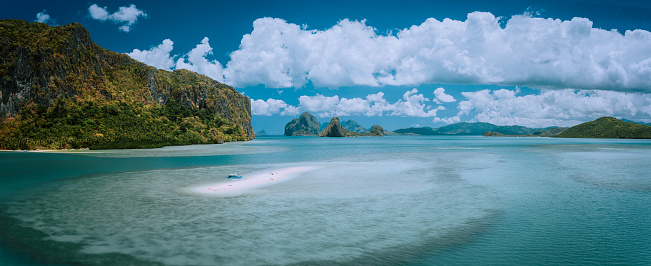 Palawan, Philippines. Aerial panoramic scenic picture of sandbar with lonely tourist boat in turquoise coastal water and cloudscape. El Nido Bacuit archipelago. Many limestone Island in background.