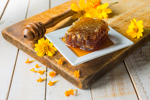 Honey composition with honeycomb, honey dipper and flowers (daisies) on old rustic wooden cutting board.