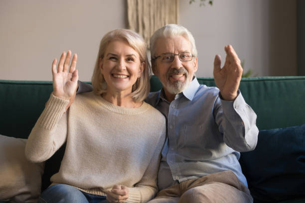 happy elderly couple looks at camera waving hands greeting friend - senior adult happiness computer looking at camera imagens e fotografias de stock