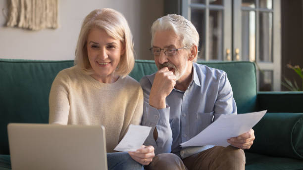 istock Elderly couple sitting on couch holding bills use computer 1158702127