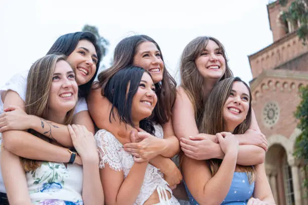 Six close girlfriends pose on their college campus for a group graduation photo. The girls in the back row wrap their arms around the girls in the front row for a hug.