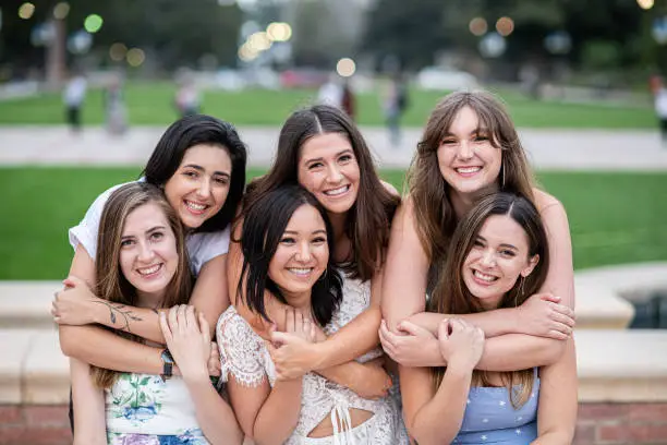 Six close girlfriends sit on the fountain edge and pose  for a group graduation photo on their college campus. The girls in the back row put their arms around the girls in the front row giving them a hug.