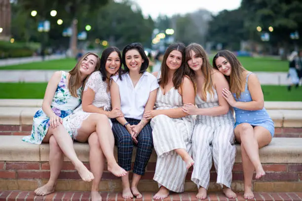 Six close girlfriends sit on the fountain edge and pose  for a group graduation photo on their college campus.
