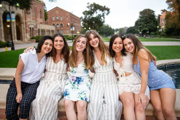 Six close girlfriends sit on the fountain edge and pose  for a group graduation photo on their college campus.