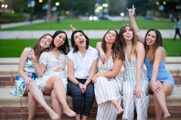 Six close girlfriends sit on the fountain edge and pose  for a group graduation photo on their college campus making silly faces.