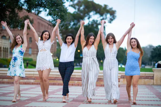 Six close girlfriends pose  for a group graduation photo. They hold each others hands up in the air as they walk towards the camera.