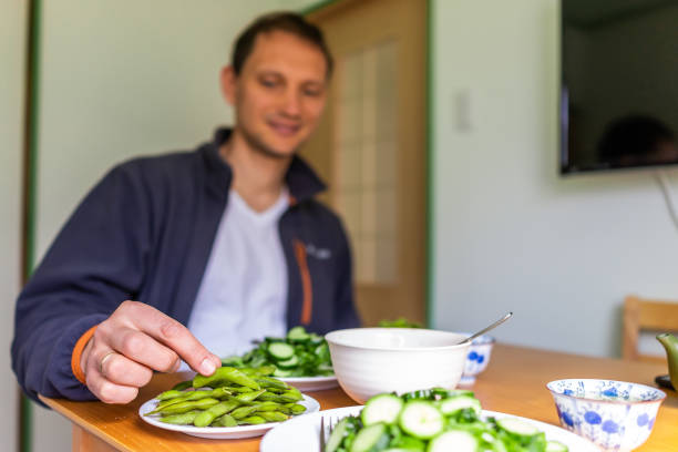 home with table and green salad dish with japanese cucumbers and mizuna greens and man trying boiled edamame soy beans - salad japanese culture japan asian culture imagens e fotografias de stock