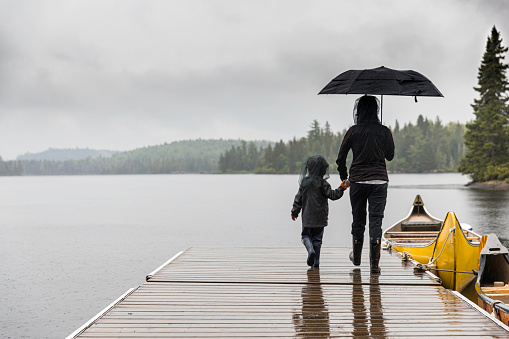 A mother and her son are holding hands on the dock by the lake in summer.  It is a rainy day and they are hiding under an umbrella. They are both wearing a mosquito netting hat to avoid mosquito bites.