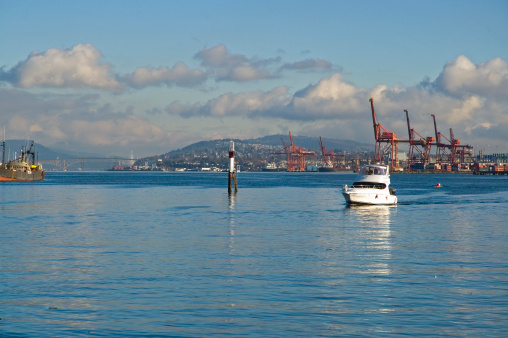 Photo shows several large Port of Vancouver cranes and docks in the background.  In the far distance to the east, you can see Workers Memorial Bridge and Burnaby Mountain where Simon Fraser University is located.  In the foreground you can see a medium sized recreational power boat sailing in Vancouver's Coal Harbour.