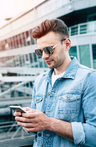 surfing in net. modern smiling handsome trendy man in sunglasses and jeans wear is using a phone outdoors in city. - surfing wireless vertical outdoors lifestyles imagens e fotografias de stock